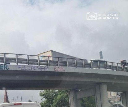Na moście widoczny jest ciężarówka oraz graffiti z napisem „WELCUM 2UER” na barierce. W tle chmury.