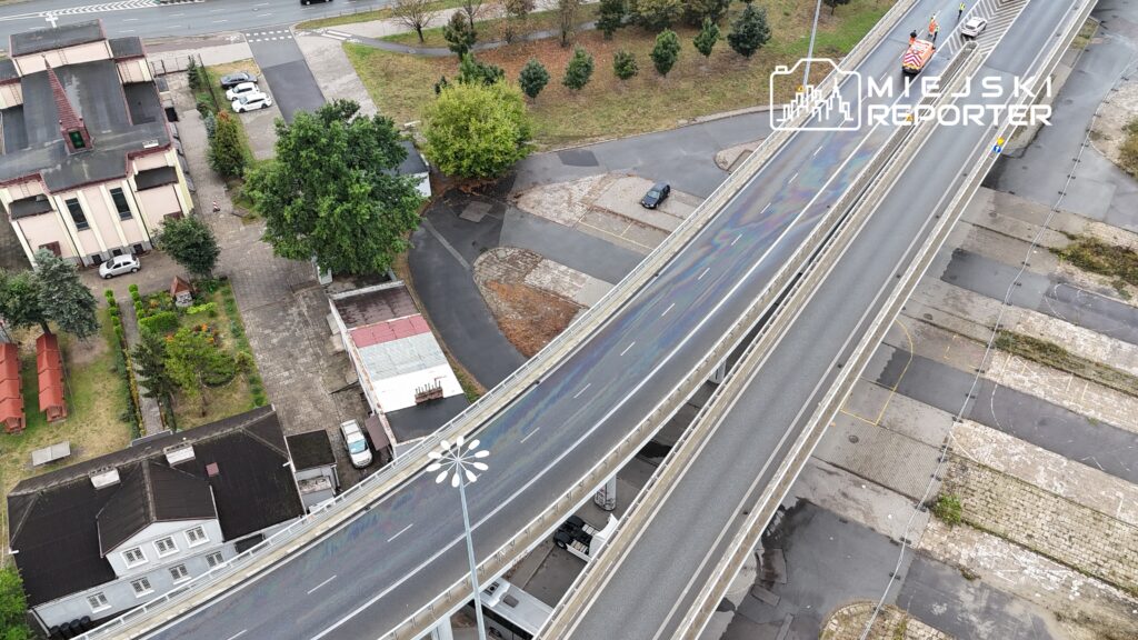 Kierowcy poruszają się po ruchliwej drodze, obok nowoczesnego wiaduktu i terenów zielonych.