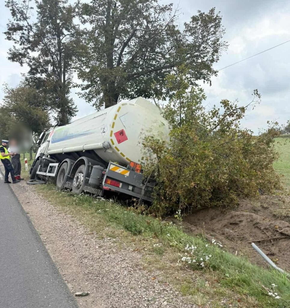 Cysterna przewrócona na poboczu drogi, obok stojący mężczyzna w kamizelce odblaskowej. W tle drzewa i pole.