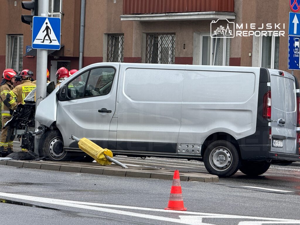 Samochód dostawczy z uszkodzoną przodem, zatrzymany przy latarni, obok grupy strażaków w mundurach.