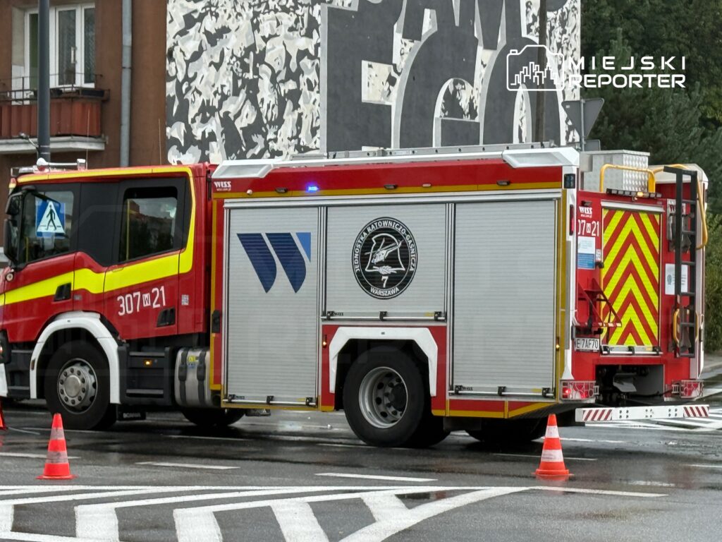 Czerwony wóz strażacki z oznaczeniami na boku stoi na skrzyżowaniu, otoczony pachołkami drogowymi. W tle widoczny mural na ścianie budynku.