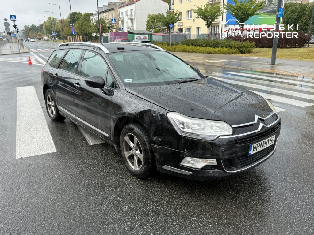 Czarny samochód osobowy zaparkowany na przejściu dla pieszych w deszczową pogodę, w tle widoczne ulice i budynki.