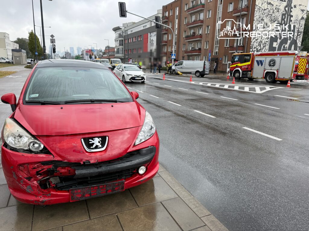 Czerwony samochód osobowy z uszkodzoną przednią częścią stoi na chodniku, obok widać akcję służb ratunkowych.
