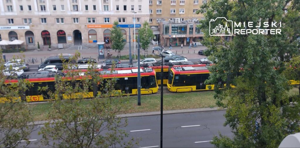 Tramwaje w kolorze żółto-czerwonym stoją na torach wzdłuż ruchliwej ulicy, obok parku z drzewami.