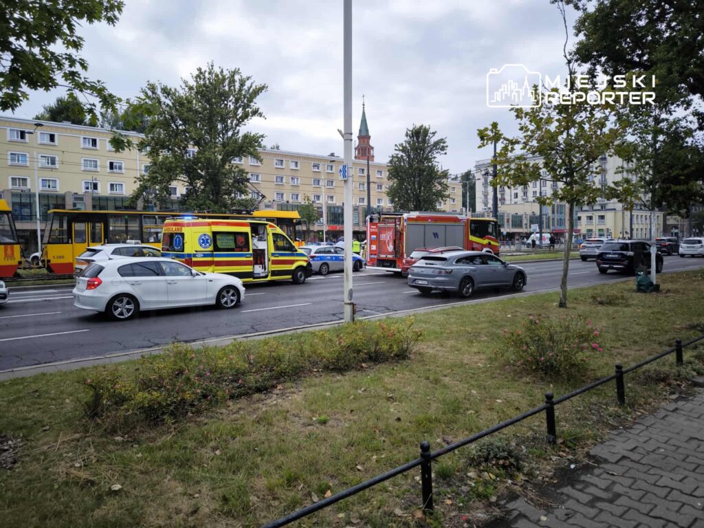 Ambulans i pojazdy straży pożarnej zatrzymane na ruchliwej ulicy, obok tramwaju i budynków mieszkalnych.
