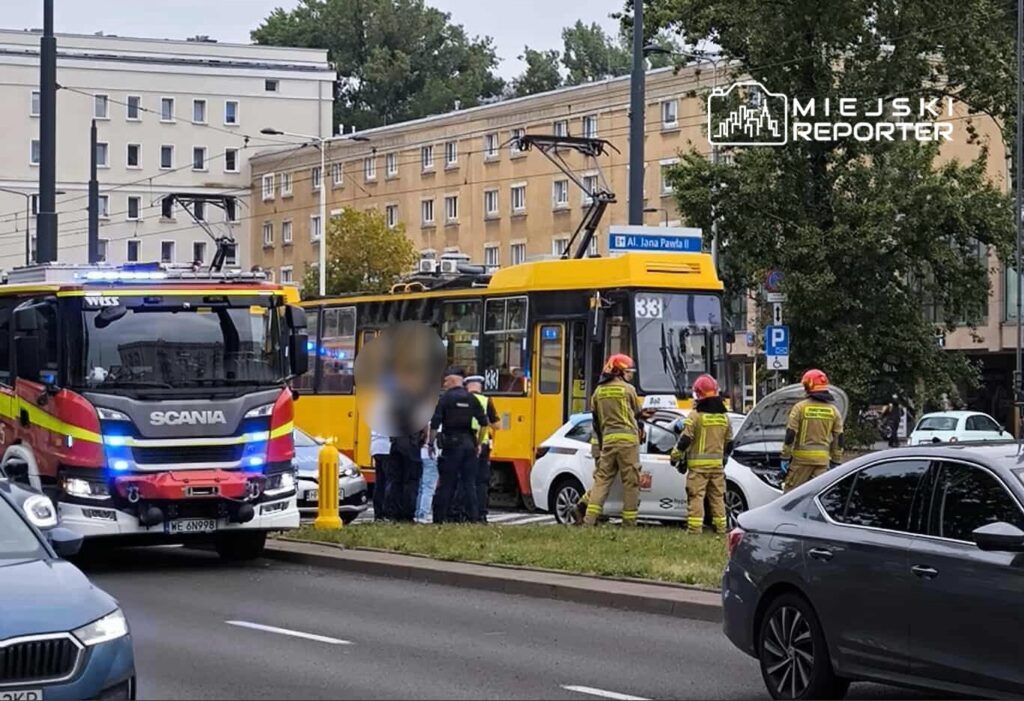 Strażacy w kaskach rozmawiają z policjantami obok uszkodzonego samochodu na ulicy, obok tramwaju.