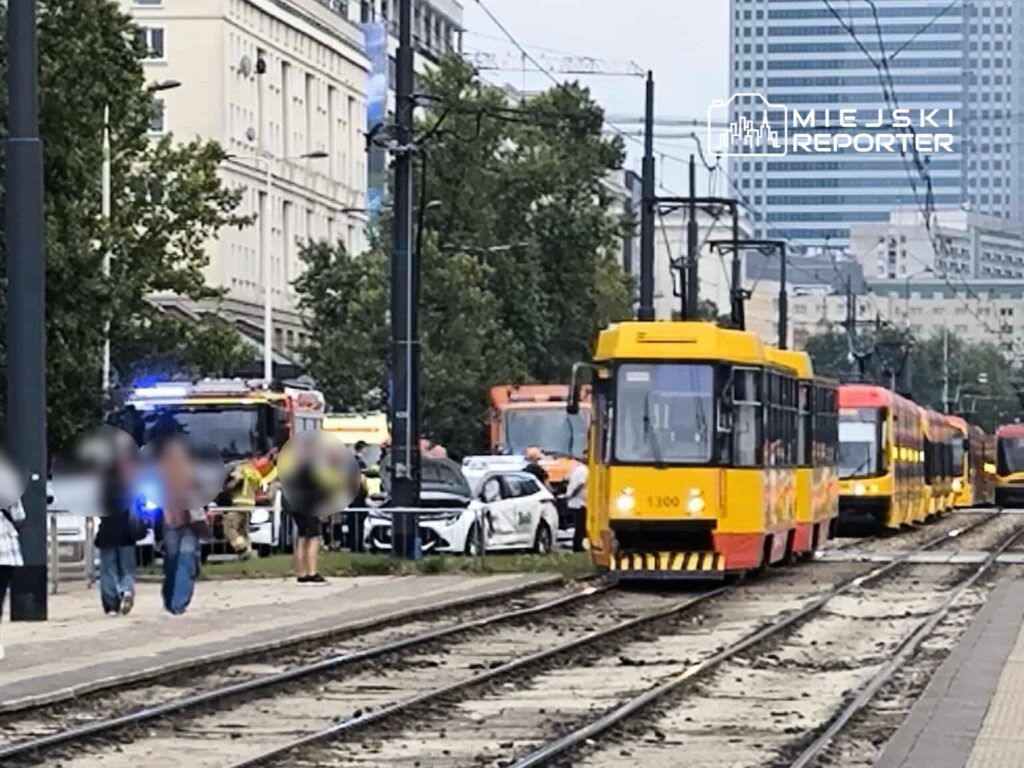 Tramwaj w kolorze żółtym zatrzymany na torach, obok grupy osób i wozów strażackich w miejskim otoczeniu.