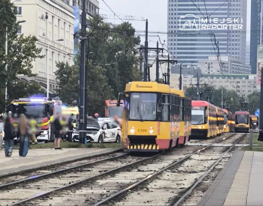 Tramwaj w żółto-czerwonym kolorze porusza się po torach, obok stoją radiowozy i pojazdy straży pożarnej.