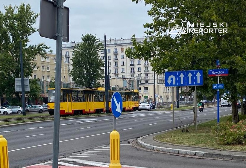 Tramwaj w żółto-czarnych barwach przejeżdża obok znaku drogowym w kierunku Nowolipia, otoczony zielenią i budynkami.
