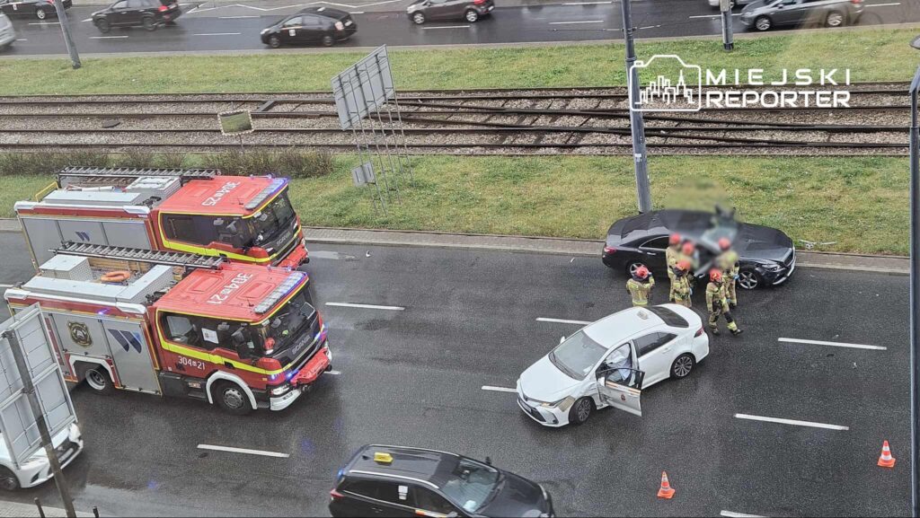 Strażacy w kaskach rozmawiają obok uszkodzonego białego samochodu na ruchliwej drodze, obok dwóch wozów strażackich.