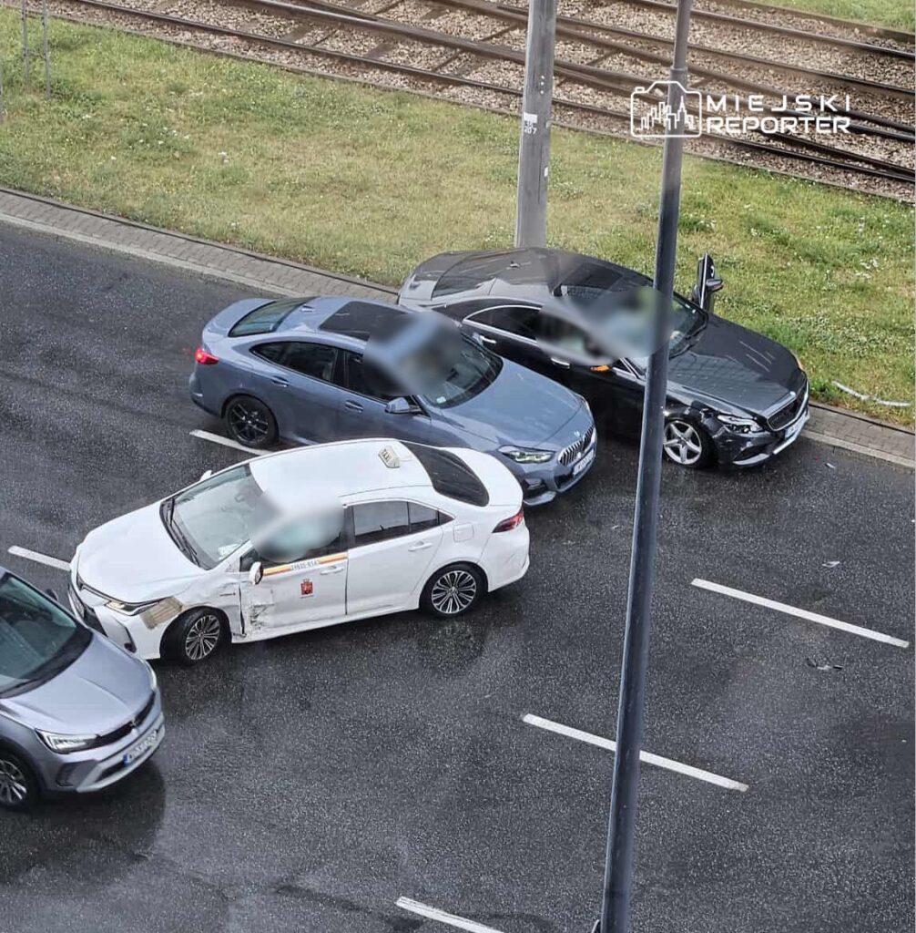 Zderzenie trzech samochodów osobowych na ruchliwej ulicy, w tym taksówki z uszkodzoną boczną częścią.