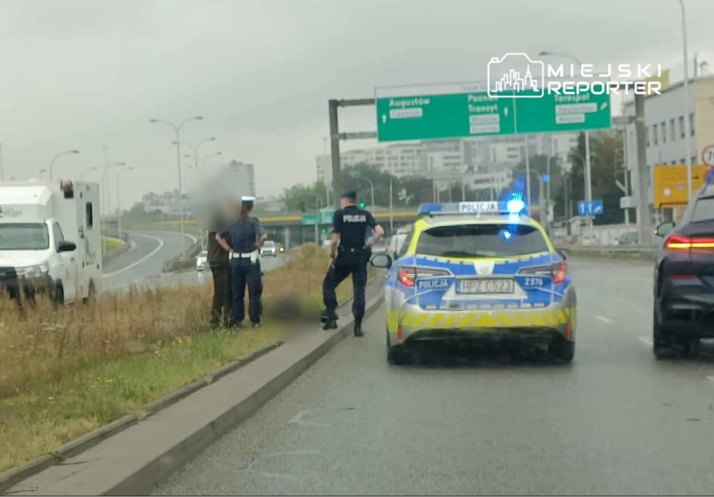 Policjanci rozmawiają przy poboczu drogi, obok radiowozu z włączonymi sygnałami świetlnymi.