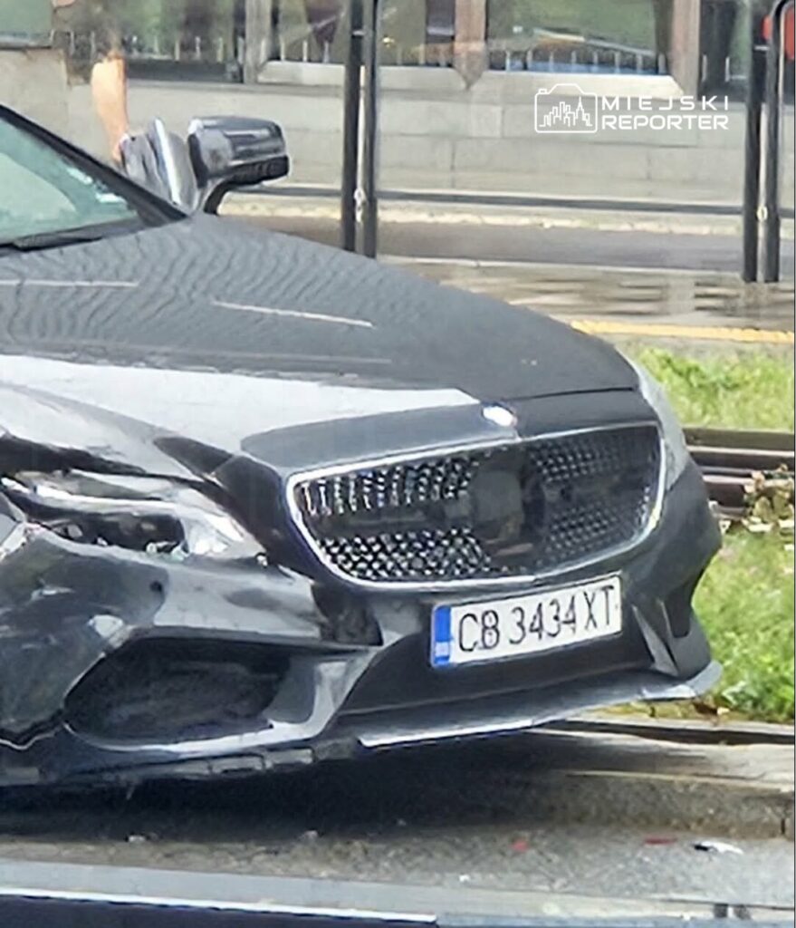 Czarny samochód osobowy z uszkodzeniami przód stoi na ulicy obok przystanku komunikacyjnego.