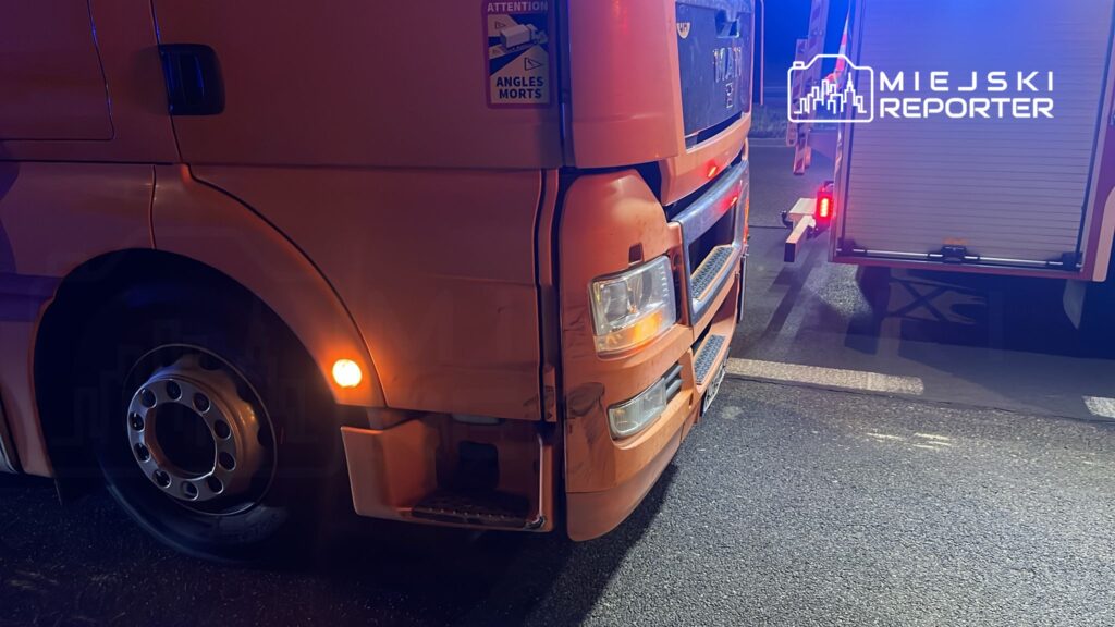 Pomarańczowy ciężarówka MAN z uszkodzoną przednią częścią stoi na drodze, obok widoczny pojazd ratunkowy.