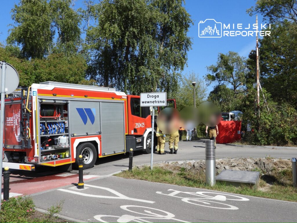 Wozem strażackim parkowanym przy drodze wewnętrznej, strażacy rozmawiają w otoczeniu zieleni.