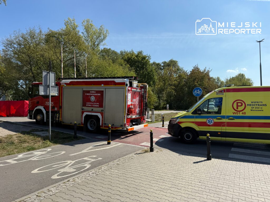 Czerwony wóz strażacki i żółta karetka stoją na drodze rowerowej w otoczeniu zieleni.