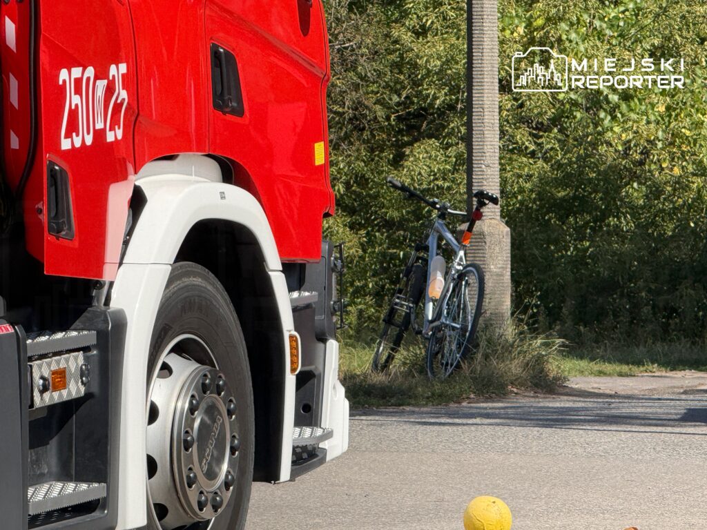 Czerwony wóz strażacki stoi na drodze obok porzuconego roweru opleanego o słup.