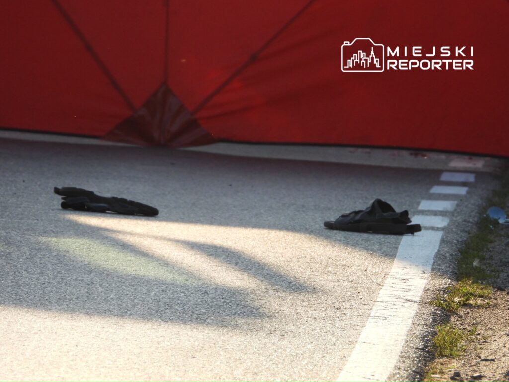 Czarne klapki leżą na asfaltowej drodze obok czerwonego namiotu, w tle widać zarys linii jezdni.