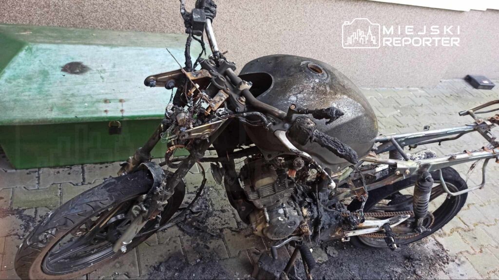 Zniszczony motocykl z popaloną ramą i silnikiem, stojący na płycie chodnikowej obok zielonego kontenera na odpady.