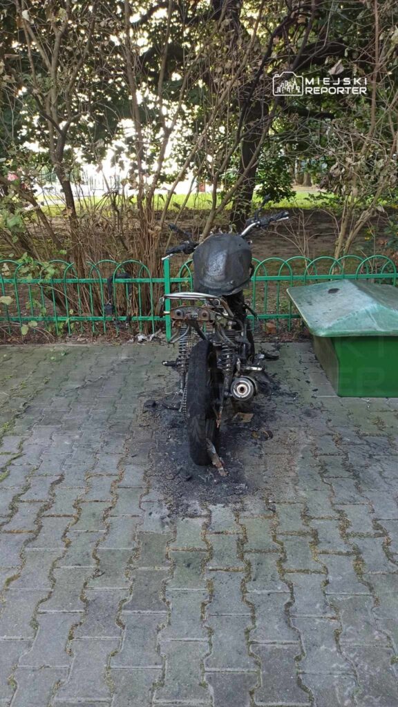 Spalony motocykl stoi na wybrukowanej powierzchni obok zielonego kosza na śmieci, w otoczeniu drzew.
