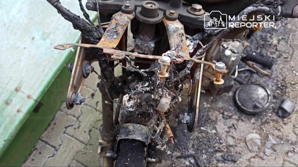Uszkodzony silnik motocykla z widocznymi śladami spalenizny i stopionymi przewodami, otoczony zniszczonymi elementami.