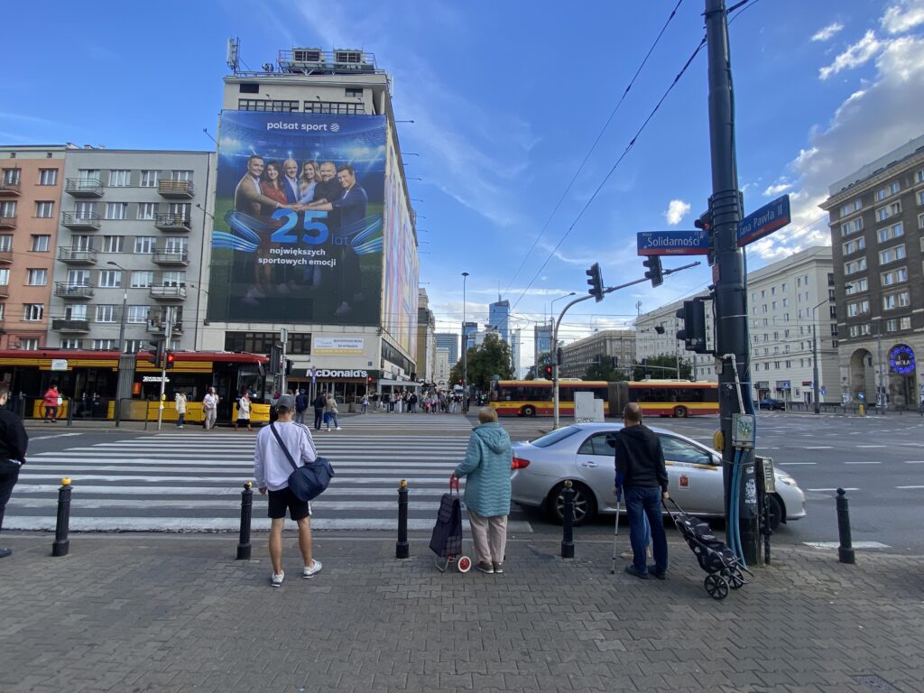 Grupa osób czeka na zielone światło na przejściu dla pieszych przed dużym billboardem reklamowym.