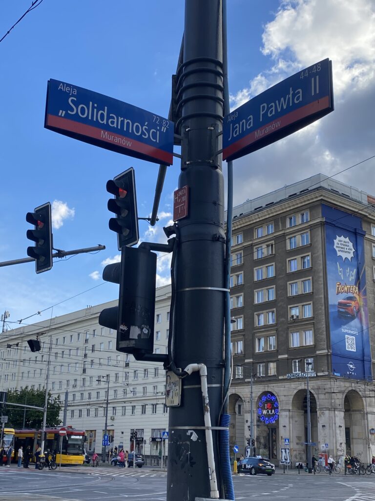 Ulica z tabliczkami "Aleja „Solidarności”" i "Aleja Jana Pawła II", w tle nowoczesny budynek z reklamami.
