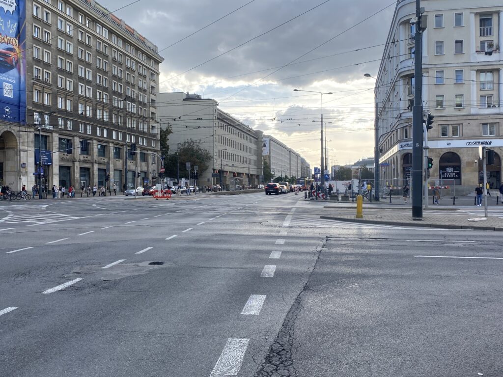 Ruchliwa ulica z nowoczesnymi budynkami w tle, samochody i piesi czekający na zielone światło.