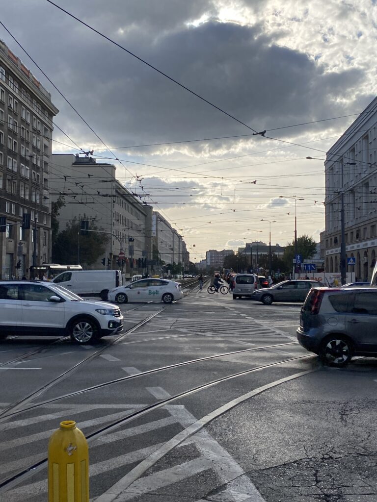 Ruchliwe skrzyżowanie uliczne z samochodami i rowerzystą, chmury na niebie oraz budynki w tle.