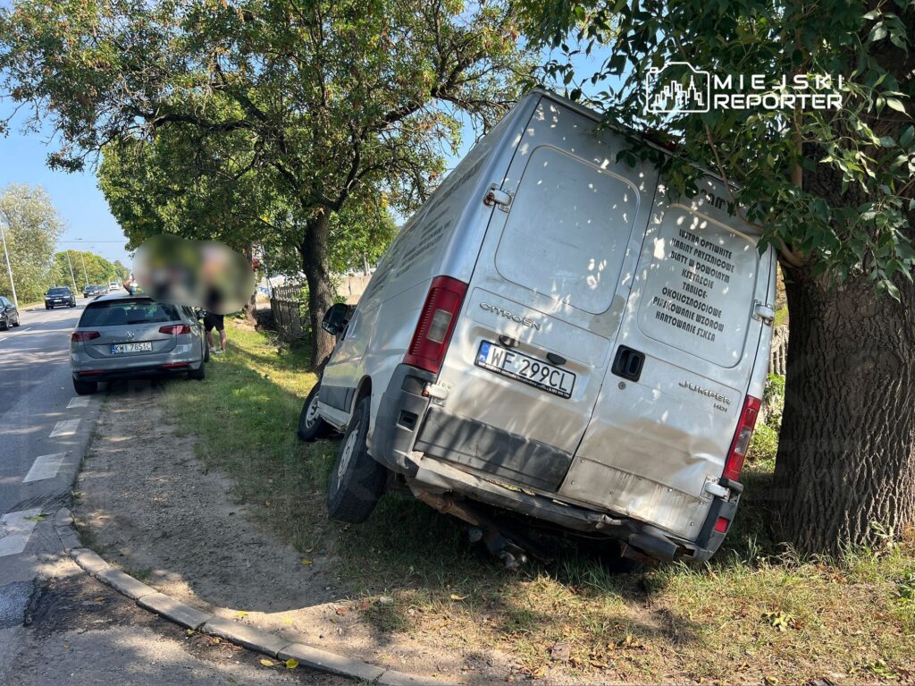 Samochód dostawczy oparty o drzewo na poboczu drogi, obok stoi osobowy samochód. W tle widoczne inne pojazdy.