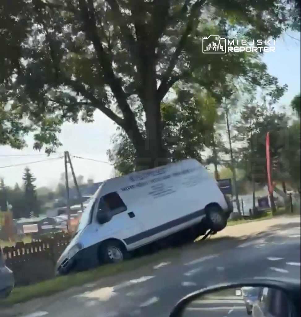 Biały furgon stoi na poboczu, z jedną stroną uniesioną nad ziemią, obok dużego drzewa i ogrodzenia.