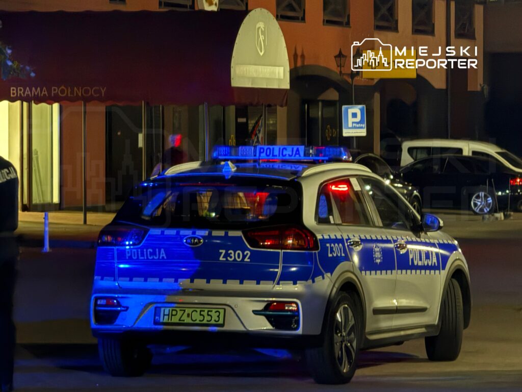 Policyjny radiowóz z niebieskimi światłami zaparkowany przed budynkiem z czerwonym daszkiem.