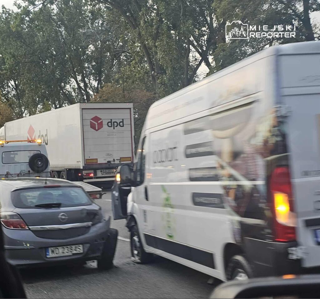 Kolizja drogowa z uszkodzonym samochodem osobowym i dostawczym w ruchu ulicznym, w tle ciężarówki.