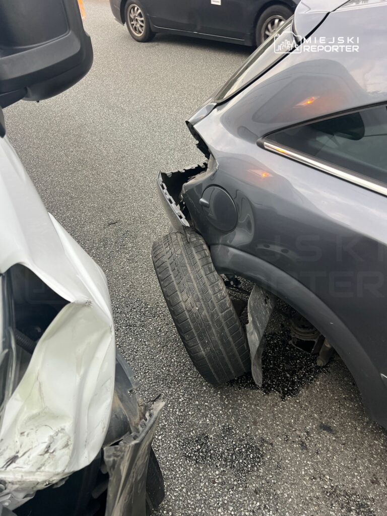 Zderzenie dwóch samochodów na jezdni, widoczna uszkodzona karoseria białego pojazdu i opona czarnego auta.