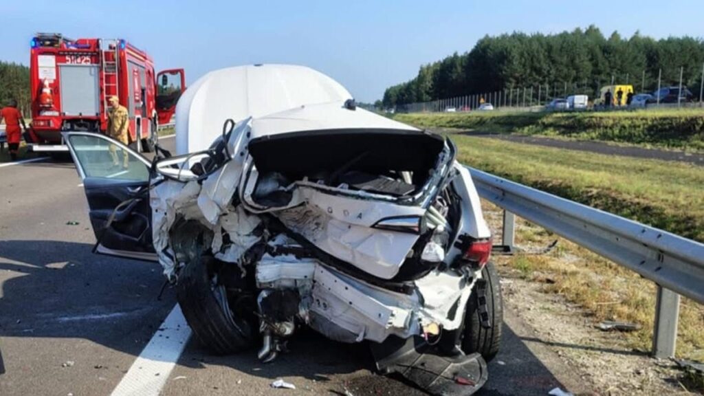 Zniszczony samochód osobowy leży na drodze, obok stoją strażacy i woz strażacki. W tle widać inne pojazdy.