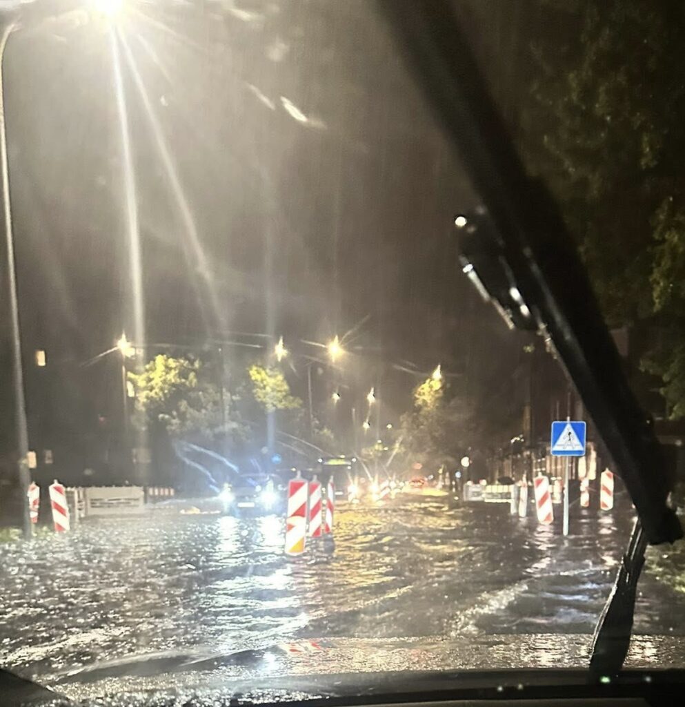 Ulica zalana wodą podczas intensywnej burzy nocą, z widocznymi znakami drogowymi i latarniami.