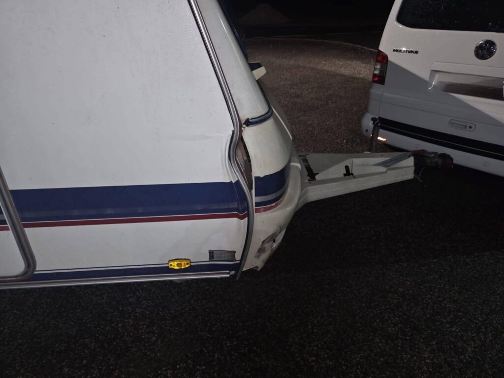 Uszkodzona przyczepa kempingowa z widoczną rysą na boku, przymocowana do samochodu osobowego na nocnym parkingu.