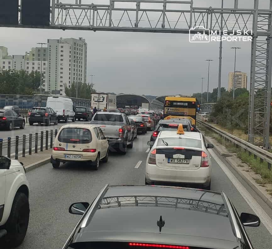Korki na drodze, wśród samochodów osobowych i autobusów, w tle nowoczesne budynki mieszkalne.