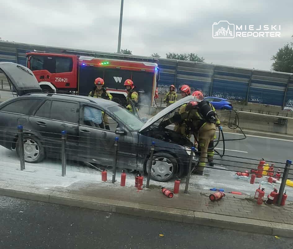 Strażacy gaszą pożar silnika samochodu na drodze, obok stoją gaśnice, a w tle widoczny wóz strażacki.