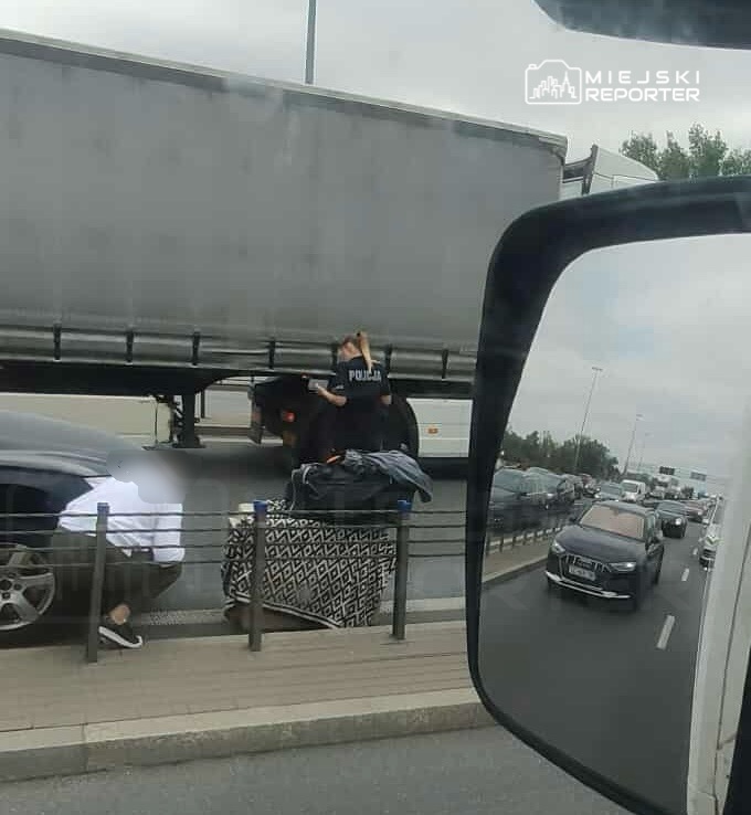 Policjant sprawdzający zawartość bagażu na chodniku obok zaparkowanego samochodu na ruchliwej drodze.