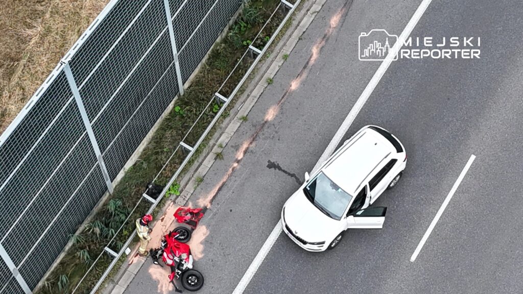 Strażak bada wypadek motocyklowy obok białego SUV-a na drodze, w tle ogrodzenie akustyczne.