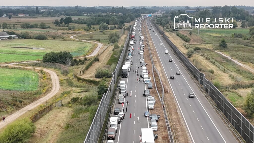 Ruch samochodowy na autostradzie z zablokowanym pasem, obok stojące pojazdy i ludzie poruszający się pieszo.