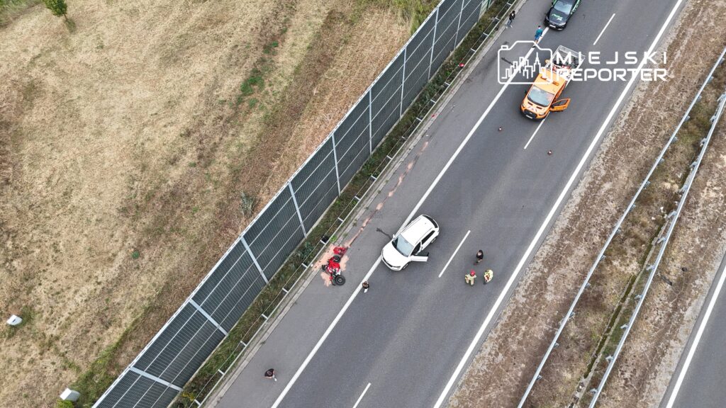Wypadek drogowy z białym samochodem na poboczu, obok leży uszkodzony motocykl, a obok stoją ratownicy i świadkowie.