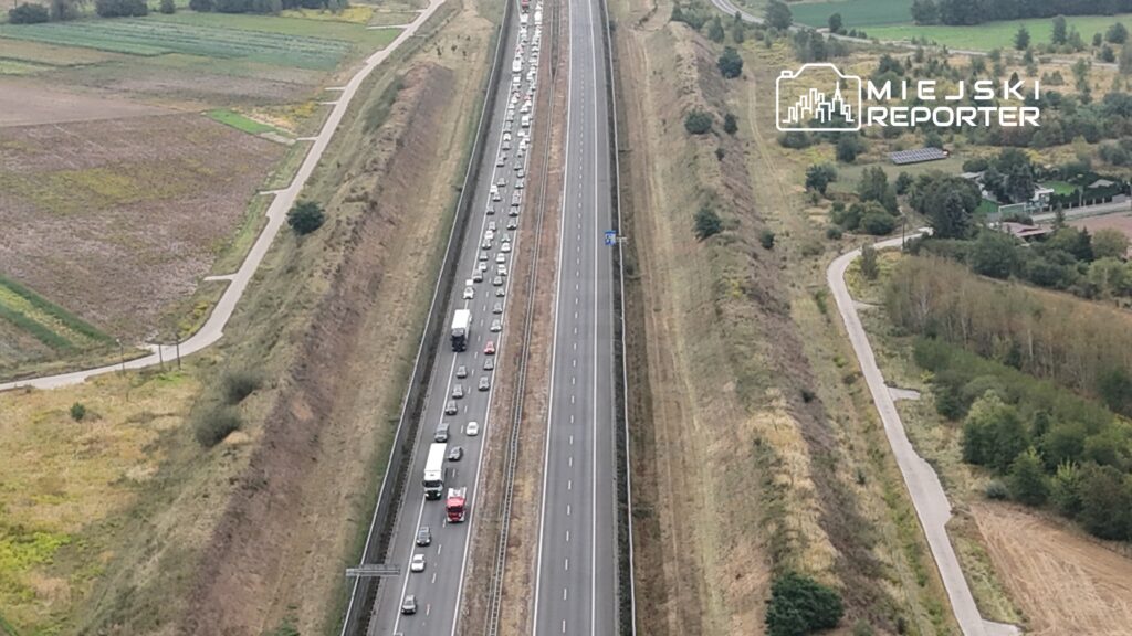 Zatłoczone pasy ruchu na autostradzie otoczonej zielonymi polami i wzgórzami.