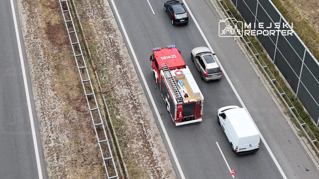 Wozidło strażackie porusza się po drodze, mijając inne samochody na pasie ruchu.