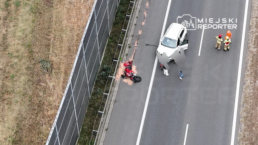 Motocykl w czerwonym kolorze leży na poboczu drogi, obok grupy osób i białego samochodu.