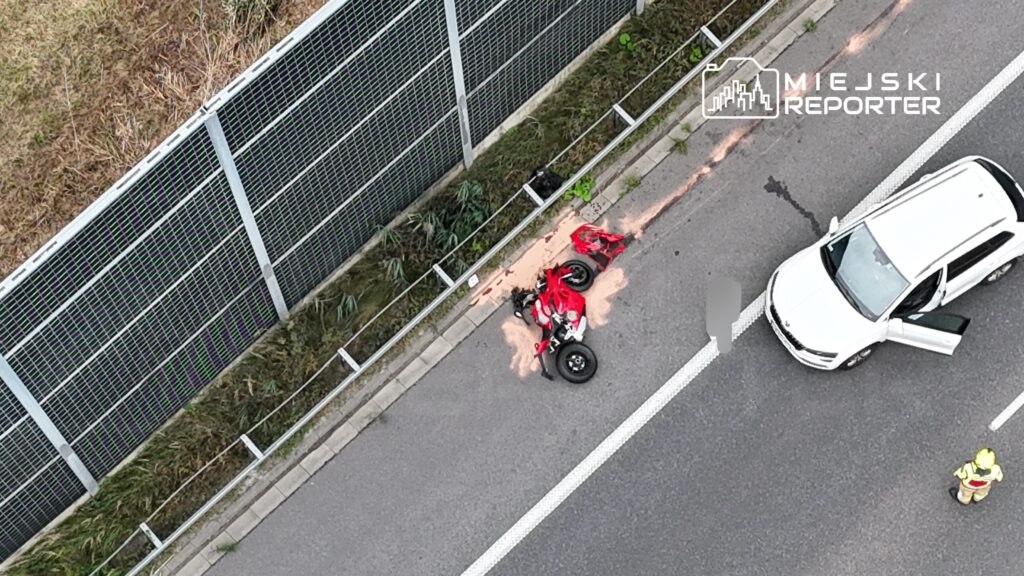 Czerwony quad leży na poboczu drogi, obok białego samochodu, a obok stoi strażak w żółtym hełmie.