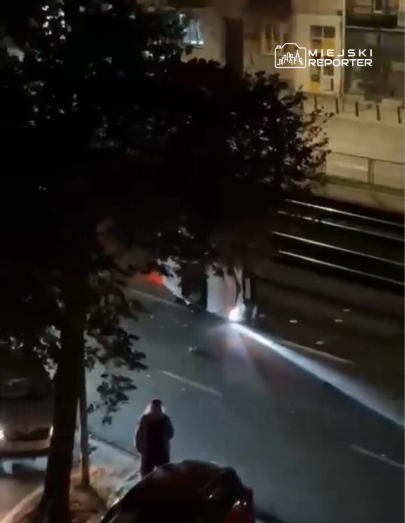 Mężczyzna w ciemnym płaszczu stoi na chodniku obok ulicy, a w tle widać światła pojazdu oświetlające drogę.