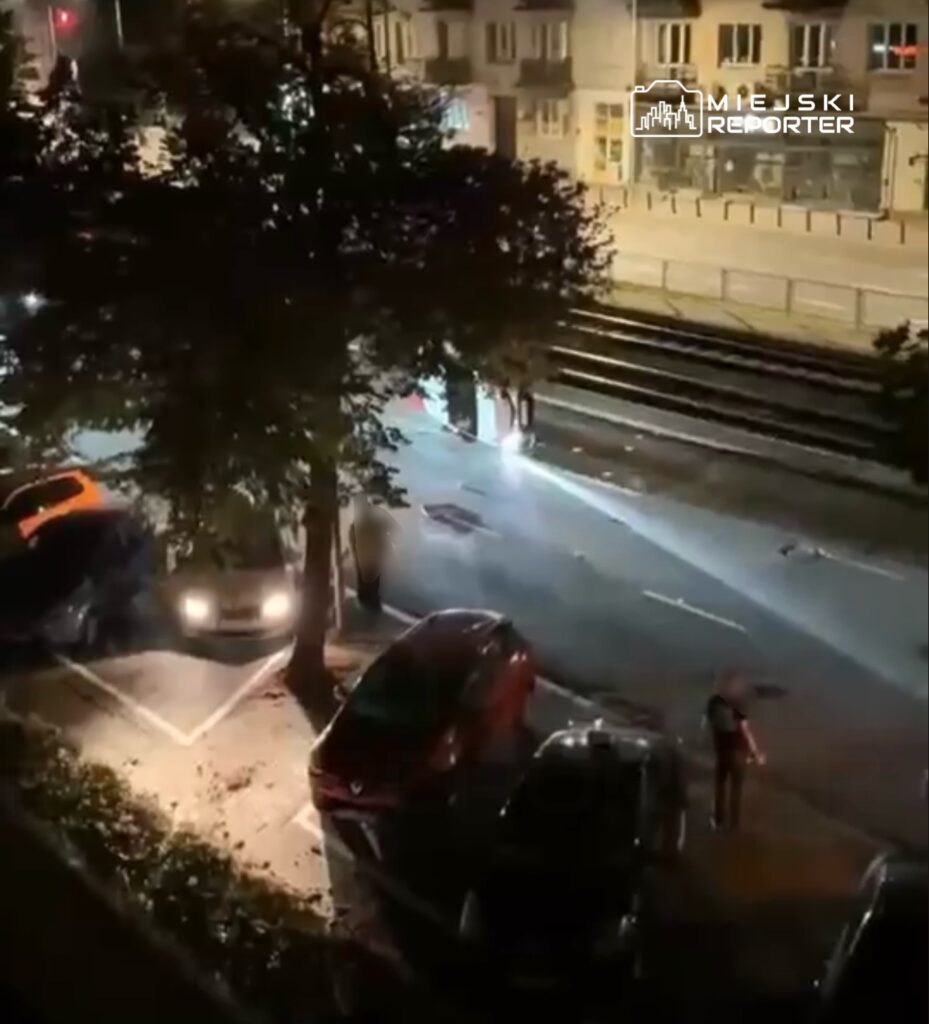 Na oświetlonej ulicy nocą, grupa osób stoi obok zaparkowanych samochodów, a w tle widać tory tramwajowe.