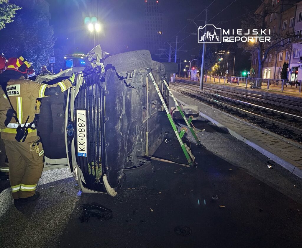 Strażak przywraca do pionu przewrócone auto na ulicy, obok torów tramwajowych, w nocy z włączonymi sygnałami.
